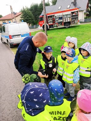Zdjęcie przedstawia policjanta, wraz z przedszkolakami. Jedno z dzieci ma na sobie czapkę policyjna oraz policyjne wyposażenie.