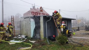Na zdjęciu widać niewielki budynek, z którego unosi się dym. Dookoła budynku pracują strażacy, jeden z nich wchodzi po drabinie na dach budynku. Widac zniszczoną w wyniku pożaru ścianę budynku.