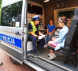 Zdjęcie przedstawia policjantkę, która siedzi w radiowozie z dziećmi i tłumaczy im przepisy ruchu drogowego.