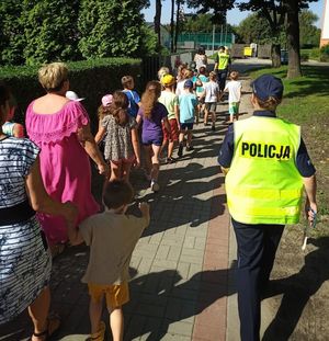 Po lewej stronie kolumna dzieci wraz z opiekunkami porusza się chodnikiem. Po prawej stronie obok dzieci w odległości kilku metrów od siebie idą dwie policjantki ubrane w kamizelki odblaskowe.