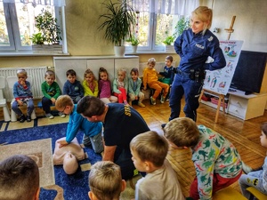 Zdjęcie przedstawia dziecko ćwiczące pierwszą pomoc na fantomie, obok stoi policjantka i strażak.