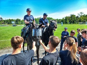 Zdjęcie przedstawia policjantów na koniach oraz zgromadzoną przed nimi młodzież.