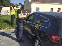 Policjant ruchu drogowego ubrany w odblaskową kamizelkę stoi przy samochodzie osobowym zatrzymanym do kontroli od strony drzwi kierowcy, drugi policjant stoi przed pojazdem.