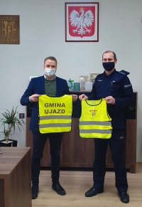 Kierownik Komisariatu Policji w Lesnicy st. asp. Arnold Skorupa oraz Burmistrz Ujazdu Hubert Ibrom trzymają w rękach małe kamizelki odblaskowe