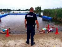 Zdjęcie przedstawia policjanta, w trakcie zajęć profilaktycznych, w tle dzieci pokonujące przeszkody nad wodą.