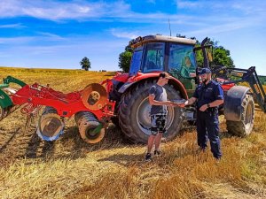 Zdjęcie przedstawia traktor podczas prac polowych,  przed nim stoi policjant oraz rolnik.
