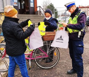 Zdjęcie przedstawia policjanta rozdającego elementy odblaskowe dwóm kobietom, obok stoją rowery.