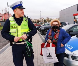 Zdjęcie przedstawia policjanta rozdającego odblaski, obok niego stoi kobieta z odblaskową torbą.