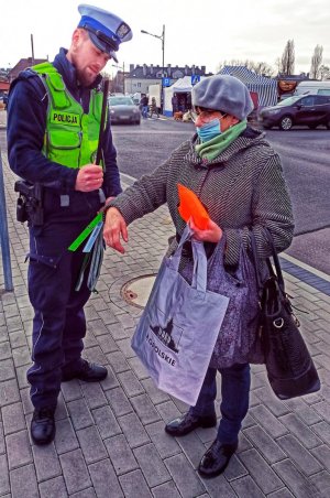 Zdjęcie przedstawia policjanta rozdającego odblaski, obok niego stoi kobieta z odblaskowa torbą i opaska naramienną.