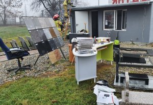 Za pierwszym planie zdjęcia widać teren przed palącym się budynkiem, gdzie na trawie  stoi biurko , na którym leżą dokumenty i przedmioty biurowe. Obok stoi krzesło biurowe, szafki i komputer. Na ziemi widać leżące kartki papieru.