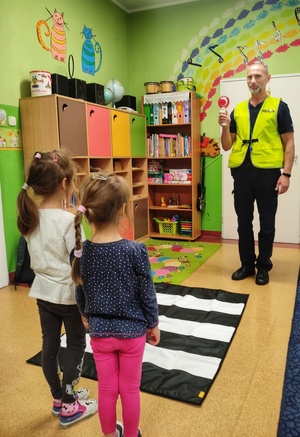 Zdjęcie przedstawia policjnata, uczącego dzieci, prawidłowego przechodzenia przez przejście dla pieszych.