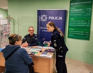 Zdjęcie przedstawia policjantów rozmawiających z seniorami na temat bezpieczeństwa. W tle budynek ZUS-u.