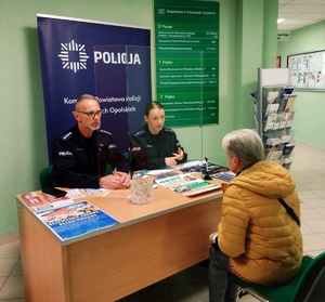 Zdjęcie przedstawia policjantów rozmawiających z seniorami na temat bezpieczeństwa. W tle budynek ZUS-u.