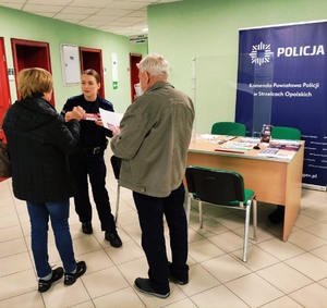 Zdjęcie przedstawia policjantkę rozmawiającą z seniorami na temat bezpieczeństwa. W tle budynek ZUS-u.