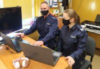 Zdjęcie przedstawia policjantów, siedzących przed komputerem, którzy za pośrednictwem Internetu prowadzą zajęcia dydaktyczne dla uczniów.