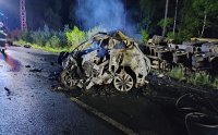 Jest ciemno, panuje noc. Na pasie jezdni położonej widać pozostałości spalonego samochodu osobowego, w tle widać leżący w rowie samochód ciężarowy.