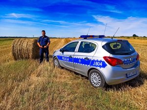 Zdjęcie przedstawia policjanta obok radiowozu, na polu, na którym trwają prace.