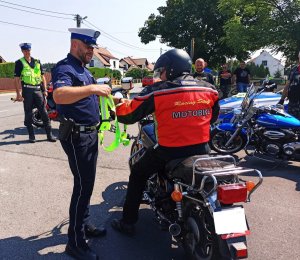 Zdjęcie przedstawia policjanta ruchu drogowego, który wręcza motocykliście szelki odblaskowe.